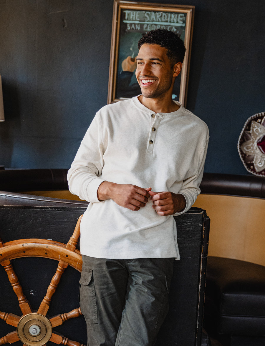 Man wearing a Waffle Knit Henley in Stone and Stretch Cargo Pant in Desert standing in a room with a wooden wheel and framed picture on the wall.
