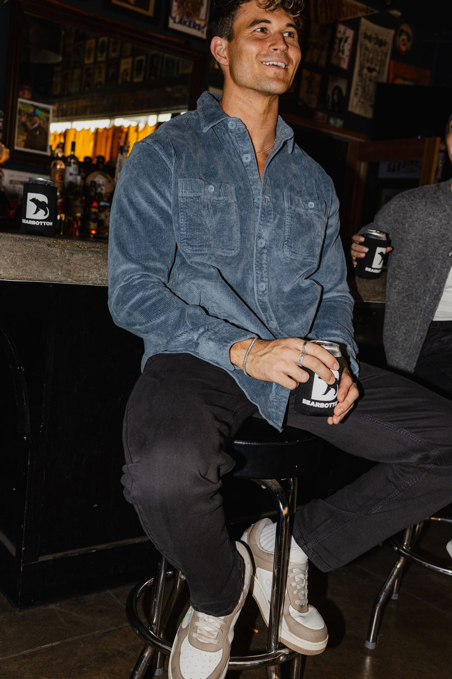 Man sitting at a bar holding a can, wearing Corduroy Workshirt in Flint and Stretch Travel Jean in Charcoal Black