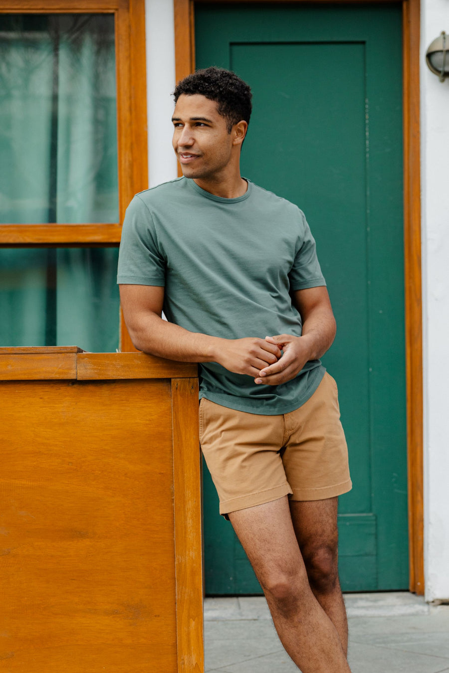 Man wearing Supima Tee in Dark Sage and stretch short in camel standing against a wooden structure with a green door in the background.