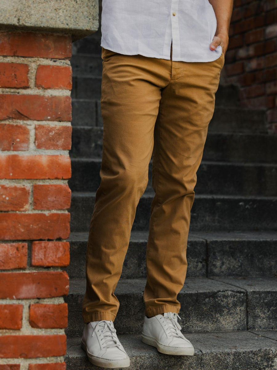 A person wearing slim-fit Stretch Chino Pants in Camel, a white button-down shirt, and white sneakers, standing on dark stone steps next to a red brick pillar.