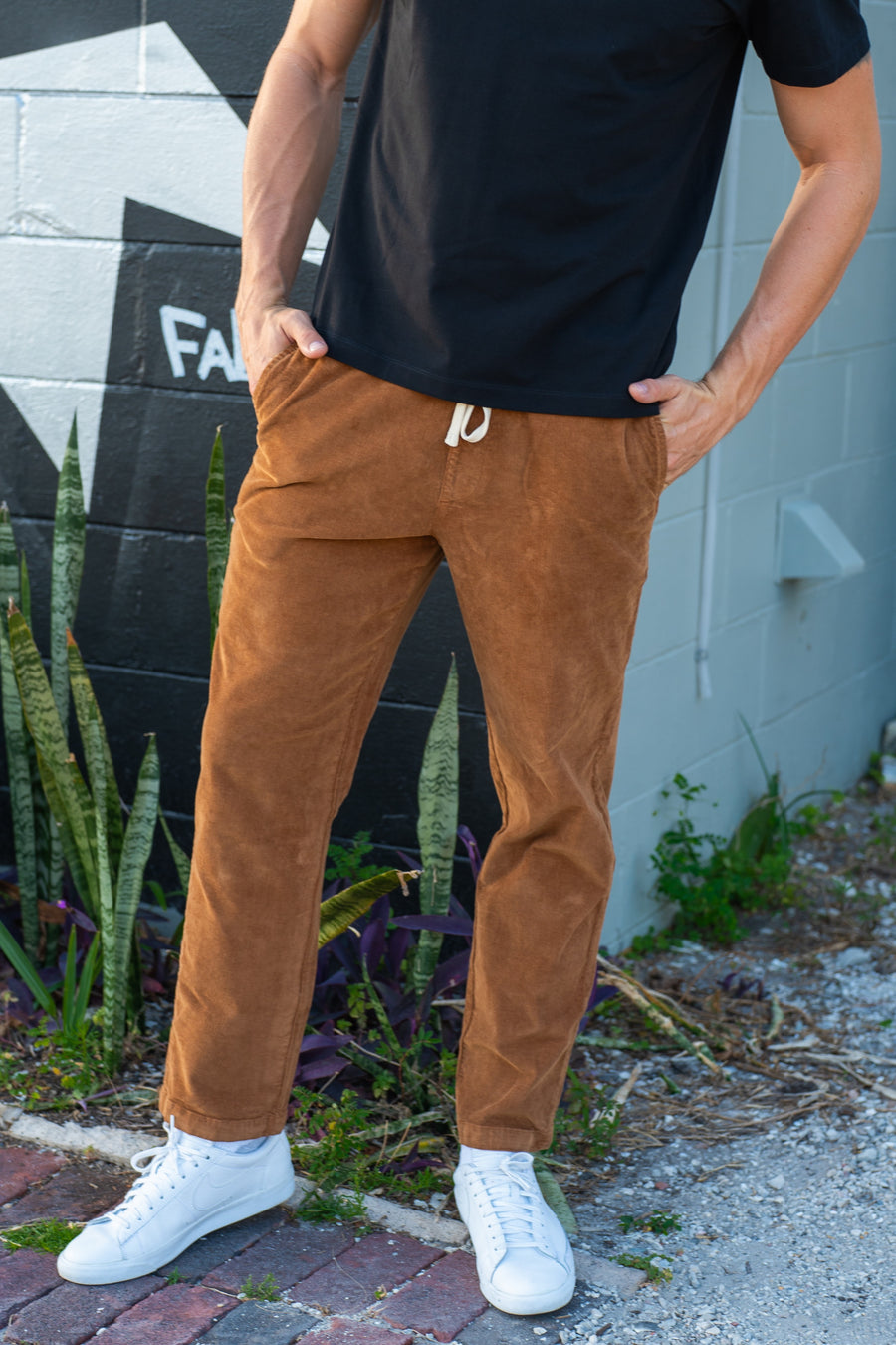 A man wearing the Corduroy Easy Pant in Dark Clay, paired with a black t-shirt and white sneakers, standing outdoors against a gray brick wall with plants.