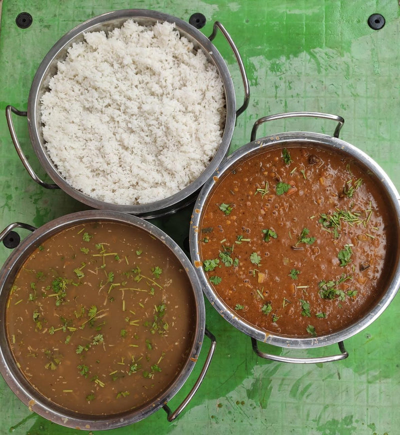 Three stainless steel bowls filled with food on a green surface: one bowl contains white rice, and the other two contain brown curries garnished with chopped green herbs.