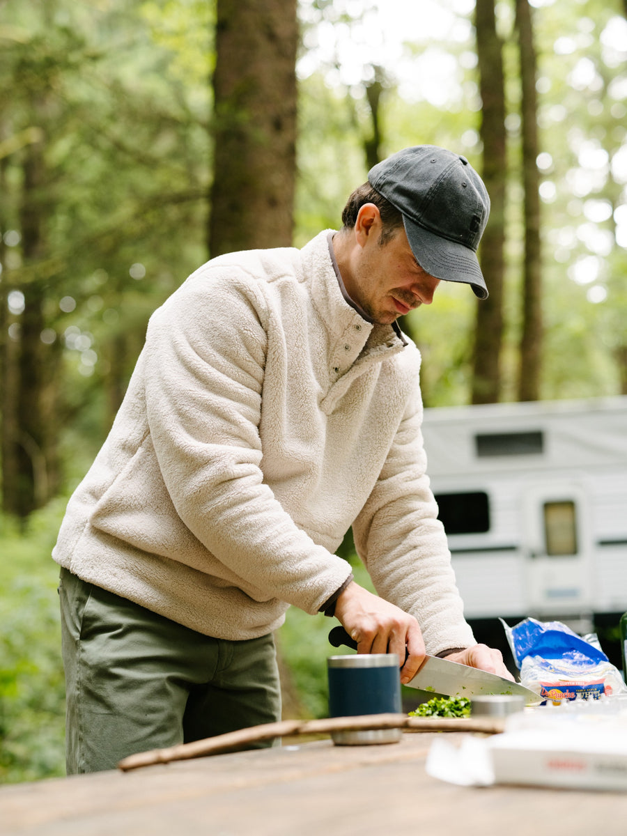 A man in the Quarter Snap Sherpa Pullover in Bone chopping food at a wooden table outdoors in a forest, with a white RV blurred in the background.