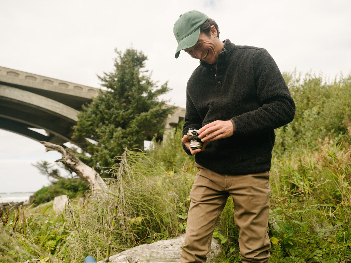 A man wearing the Quarter Snap Sherpa Pullover in Black stands outdoors in a grassy area near a bridge, holding a small camera or device.