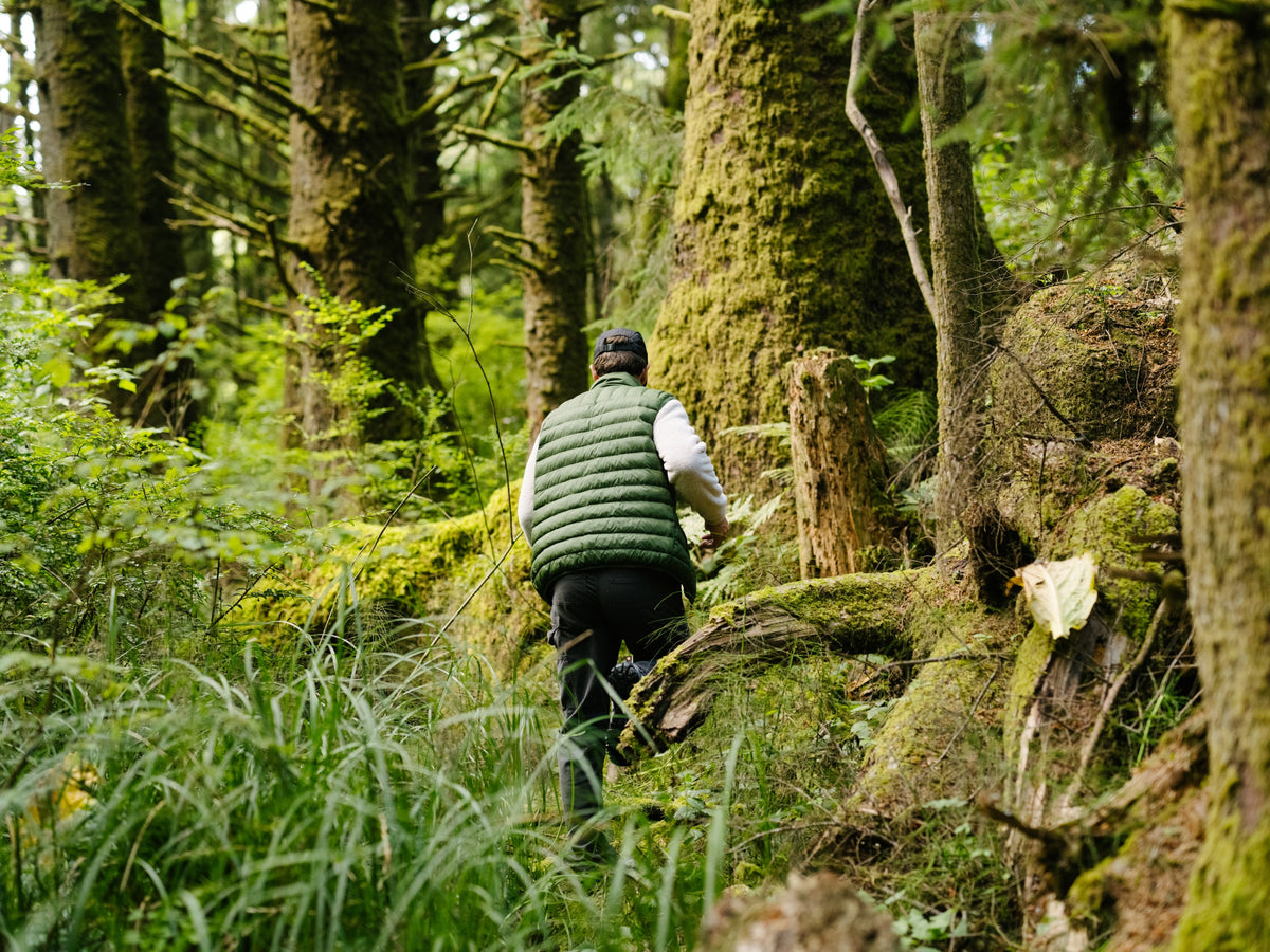 A man walking through the forest wearing the PrimaLoft Packable Vest in Pine Green.
