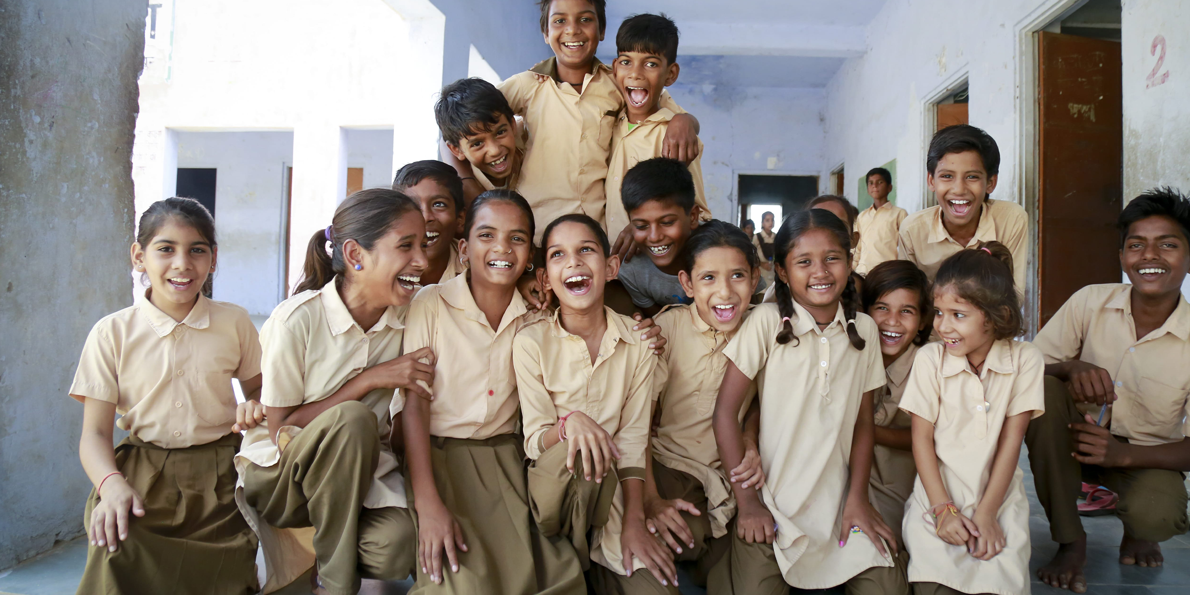 Group of children in India wearing school uniforms smiling and laughing. Text overlay states "Giving Back. 1 Item Purchased = 1 Meal Donated."