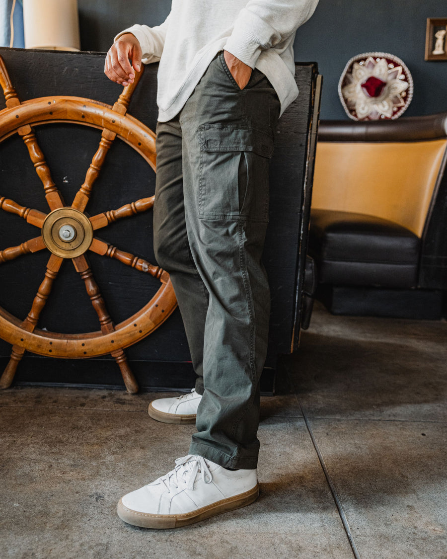 A person wearing the Military Green Stretch Cargo Pants, the Waffle Knit Henley and white canvas sneakers stands indoors next to a large wooden ship's wheel.