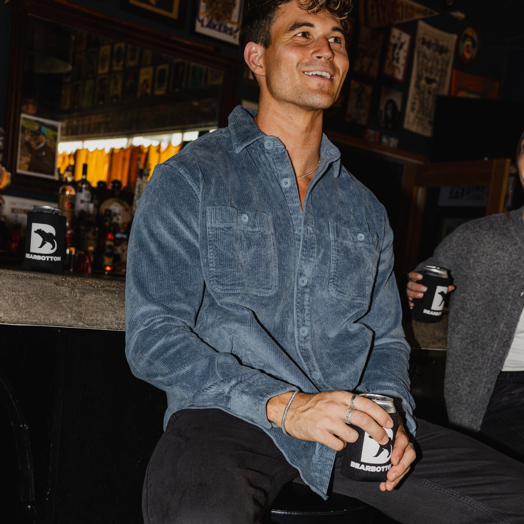 Man sitting in a bar holding a can with a Bearbottom logo, surrounded by bar decor. Man wearing Corduroy Workshirt in Flint.