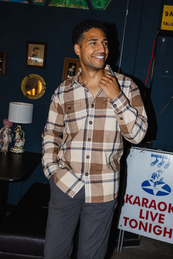 A man smiling in the Heavyweight Flannel Overshirt in Mocha Indigo standing in a dimly lit room with a blue wall, framed pictures, and a karaoke sign in the background.