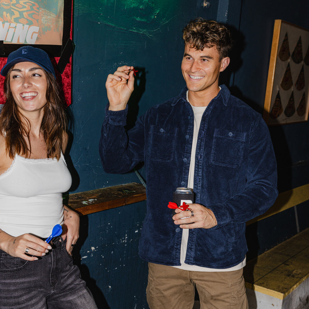 Two people at a bar, one holding a dart and smiling, with a colorful background. Man wearing Corduory Workshirt in Navy.