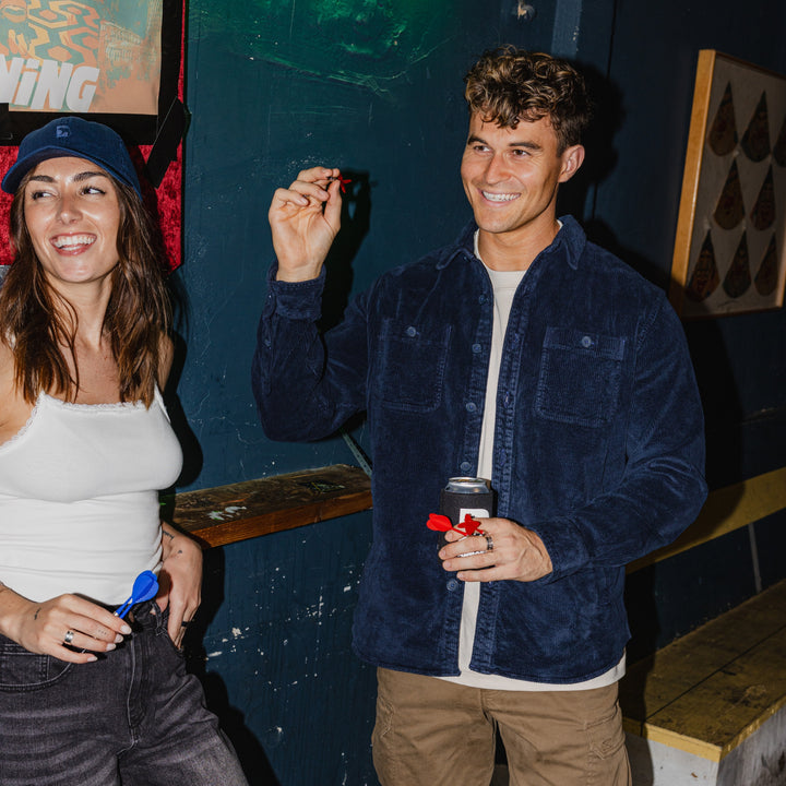 Two people at a bar, one holding a dart and smiling, with a colorful background. Man wearing Corduory Workshirt in Navy.