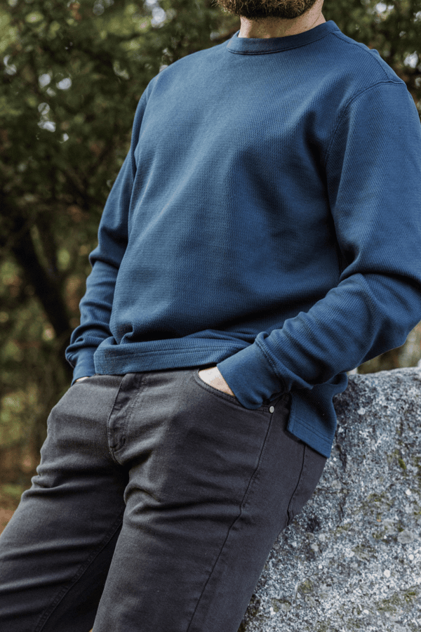 A man wearing the Thermal Knit Crew in Navy and the Stretch Travel Jeans in Charcoal Black, leaning against a large gray stone outdoors with trees in the background.