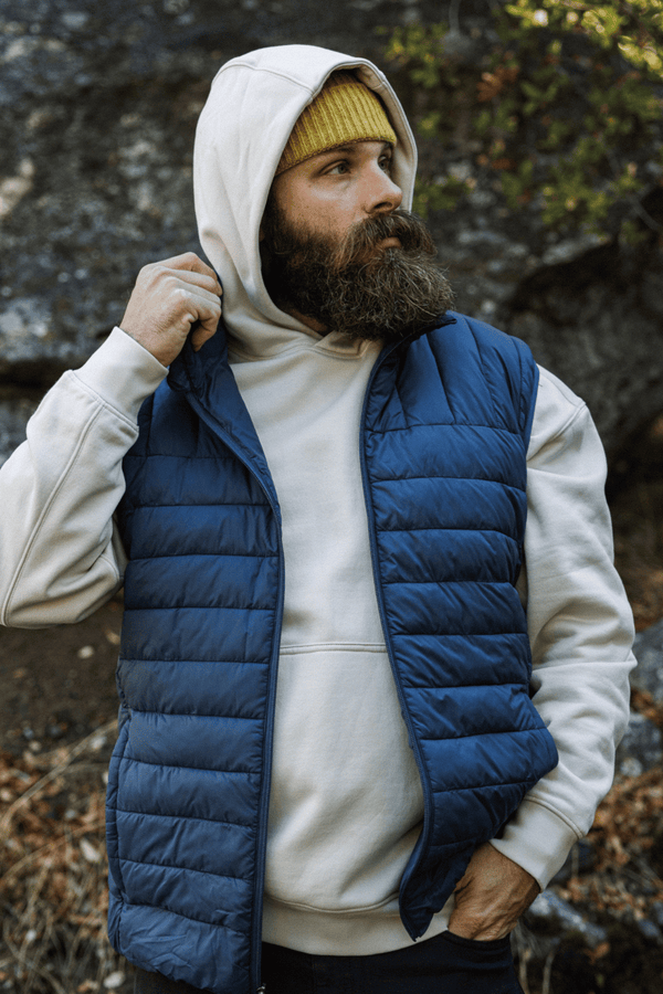 A bearded man wearing the Primaloft Packable Vest in Navy and the Core Hoodie in bone with a yellow knit beanie, standing outdoors against a rocky, wooded background.
