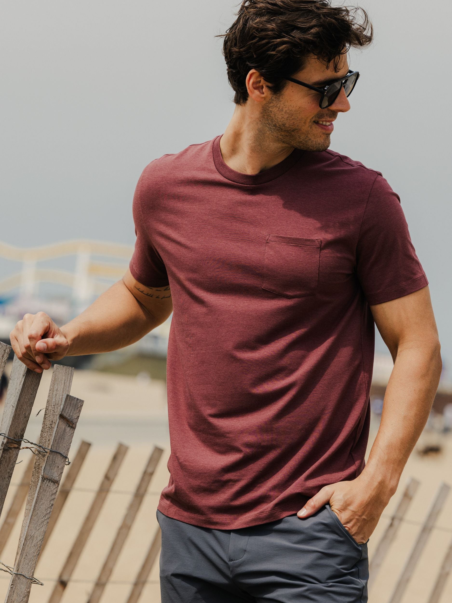 Man wearing bamboo pocket tee in deep mauve and sunglasses by a wooden fence.