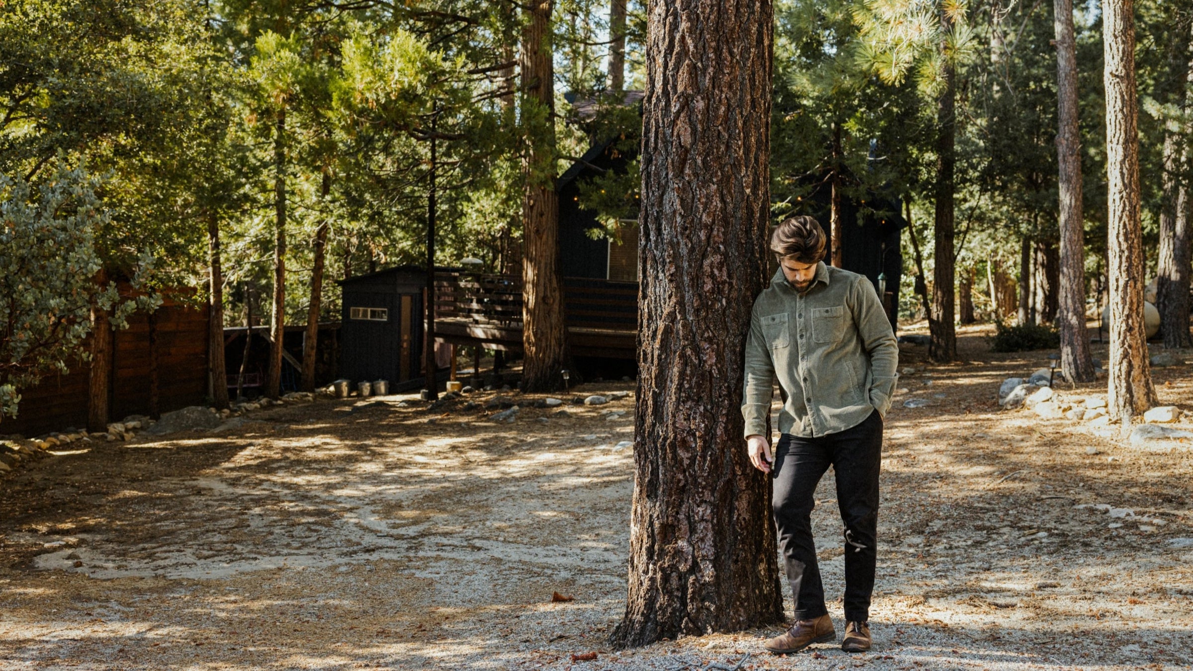 Model wearing Corduroy Workshirt Fern leaning on tree in forest in front of cabin