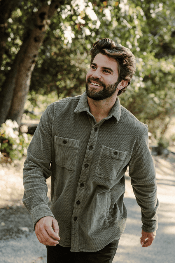 A smiling man with a beard wearing the Corduroy Workshirt in Fern standing outdoors on a sunlit path with trees in the background.