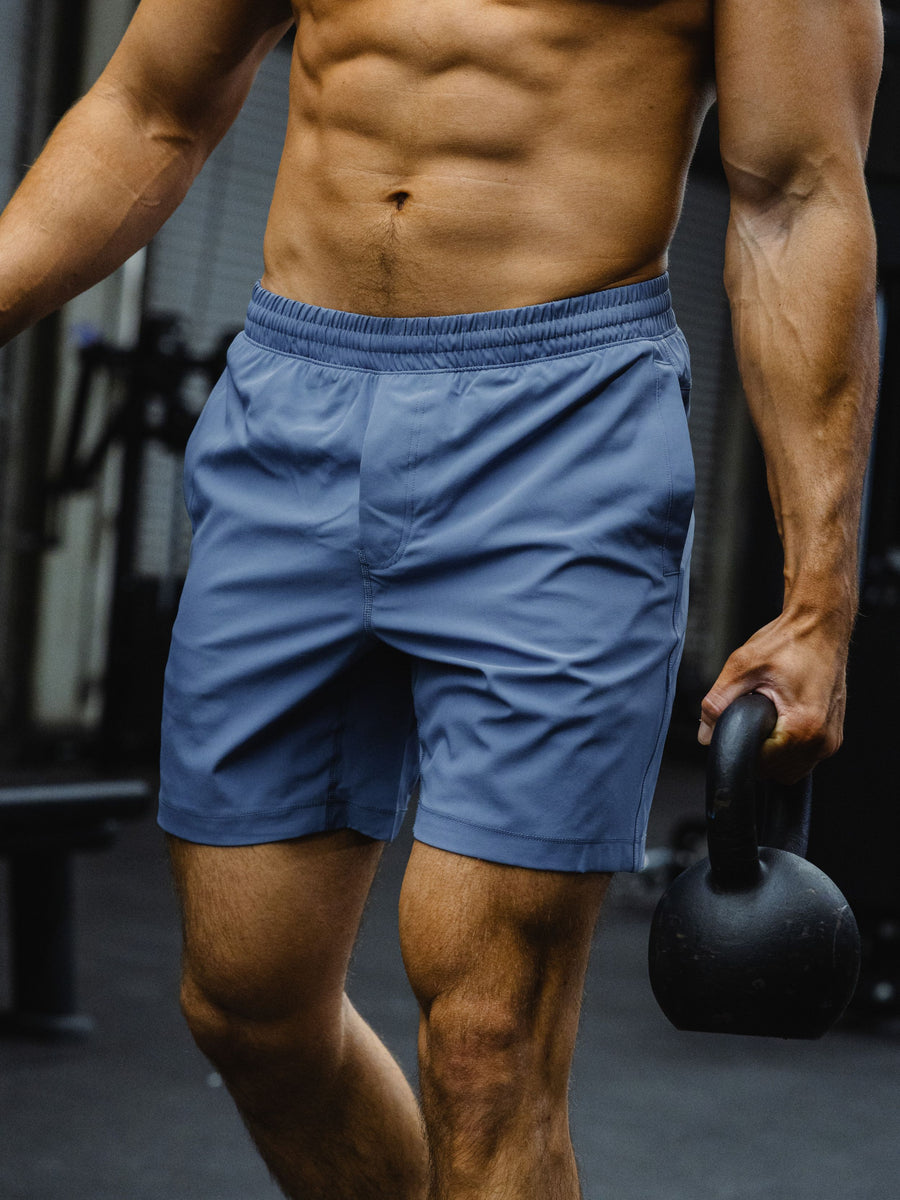 A close-up of a muscular man in a gym wearing the Drifter Short in Blue Jay and holding a black kettlebell.