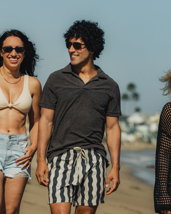 A man wears the Stretch Swim Trunk (Clearance) in Orca Wavy Stripe, 5.5" inseam, on the beach.