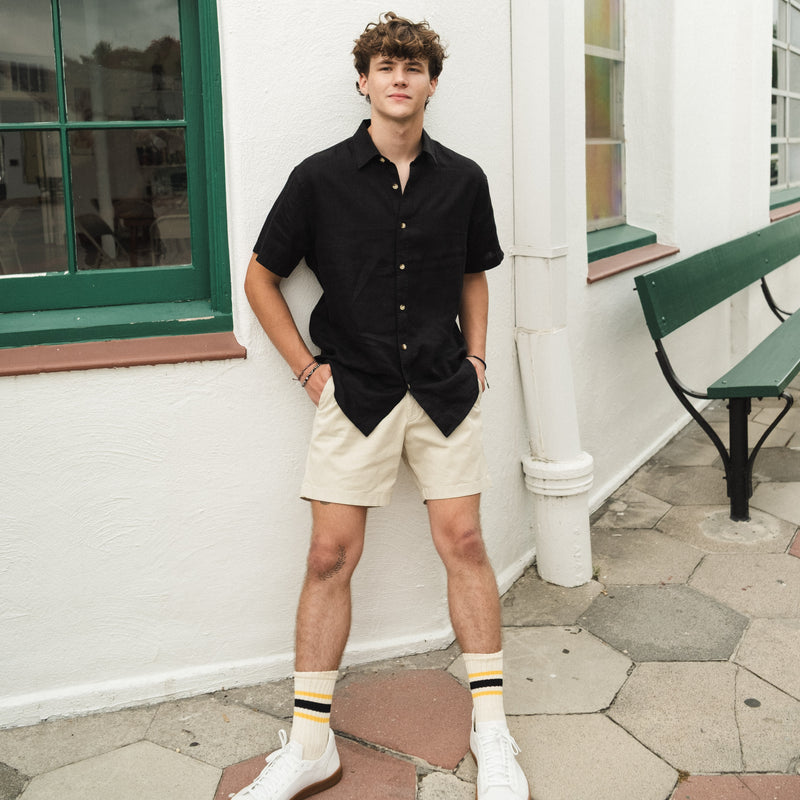 Young man linen short sleeve shirt in black and beige stretch shorts standing against a white wall with a green bench.