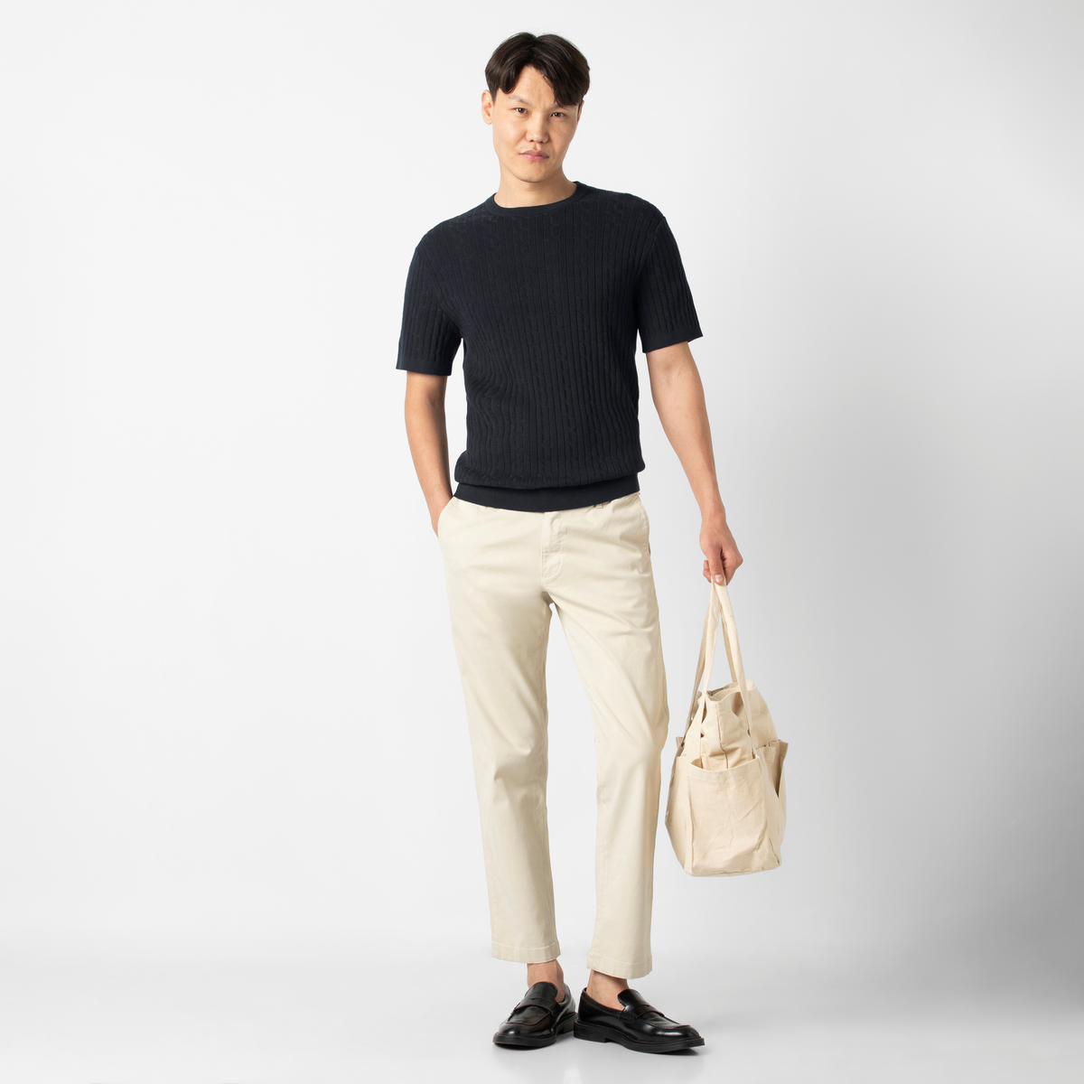 Model wearing Cable Stitch Sweater Crewneck in Black paired with Relaxed Stretch Chino Pant Stone, carrying a an off white canvas tota
