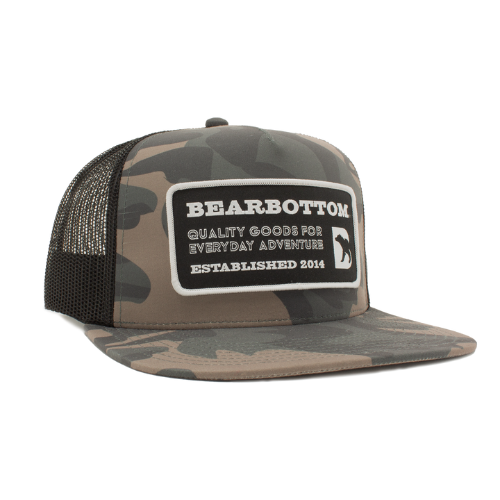 Camo Trucker Hat with camo print, flat brim, mesh back, and black patch that states BEARBOTTOM. QUALITY GOOD FOR EVERYDAY ADVENTURE. ESTABLISHED 2014.