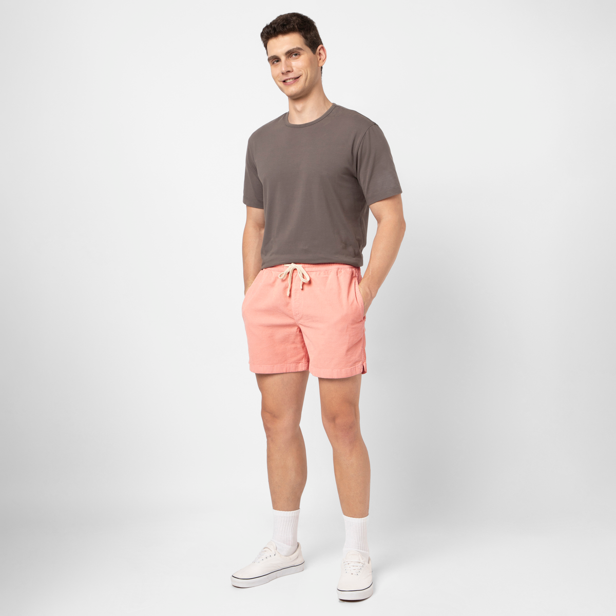 A man stands smiling against a plain white background, wearing a gray t-shirt, Corduroy Easy Short in light pink with a relaxed fit and drawstring waist, white crew socks, and white sneakers.