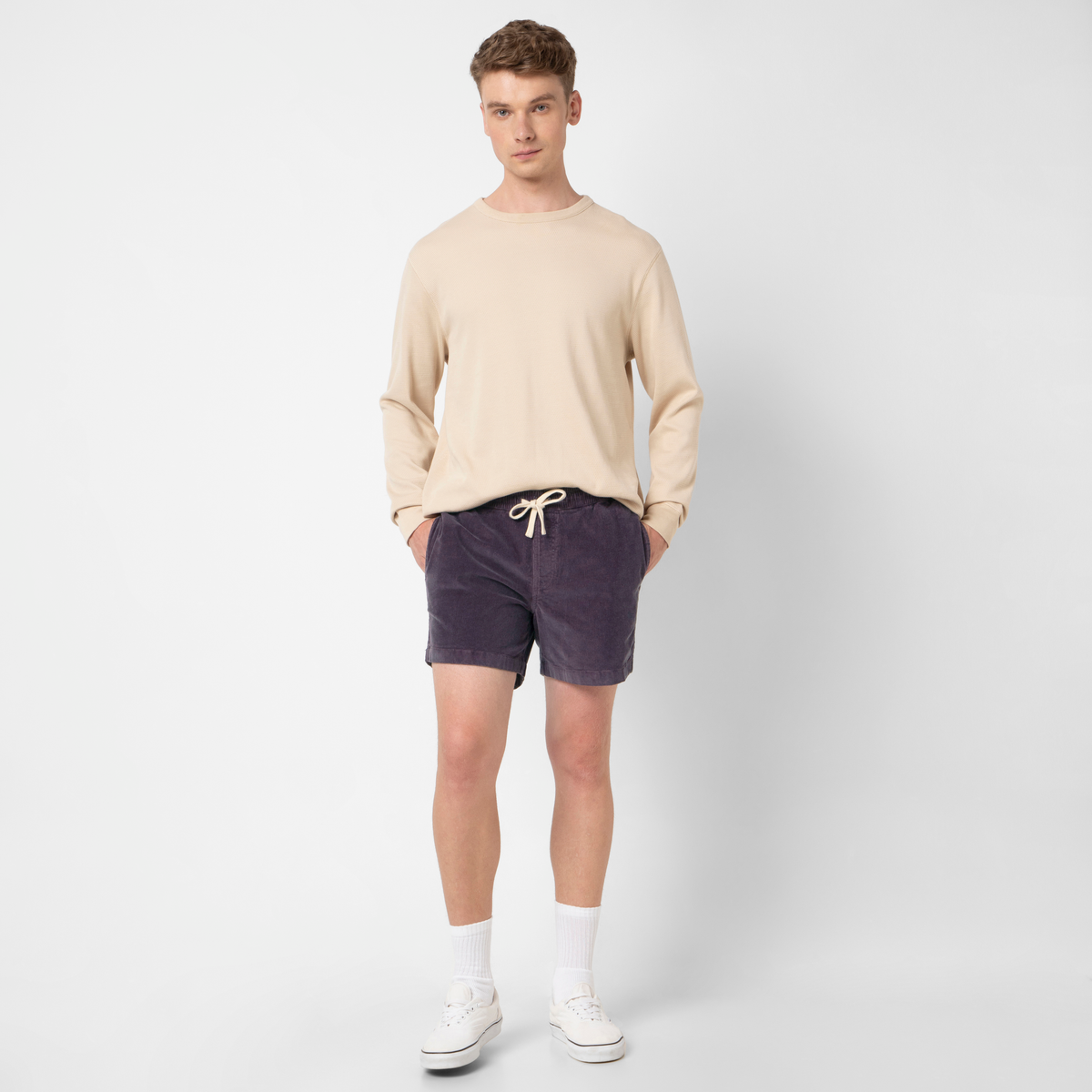 A young man stands against a plain white background wearing the Corduroy Easy Short in beige, paired with a matching long-sleeve shirt, white socks, and sneakers. His hands in his pockets complete the classic style look.