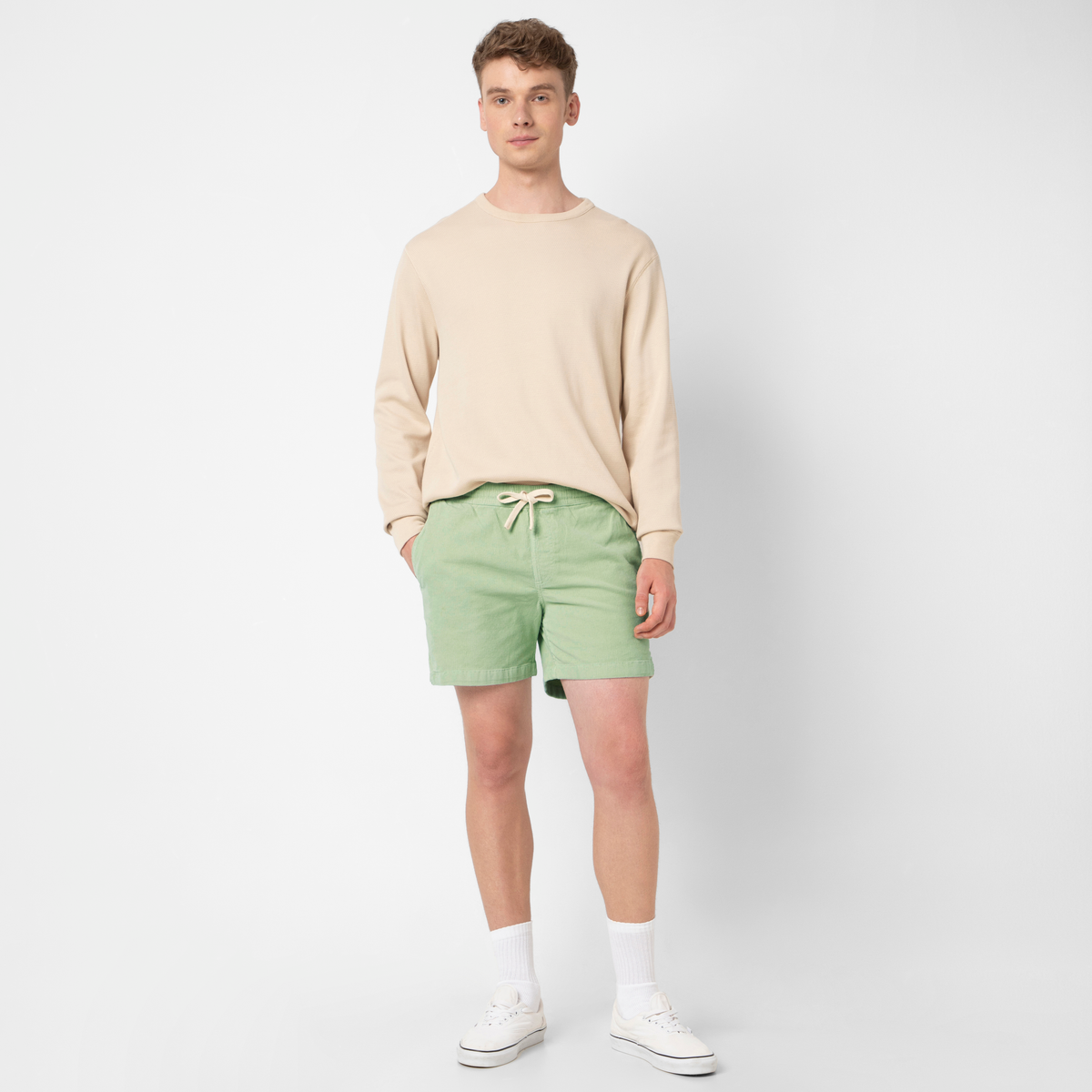 A young man faces forward, wearing Corduroy Easy Shorts in light green with a beige long-sleeve shirt, white socks, and sneakers against a plain white background.