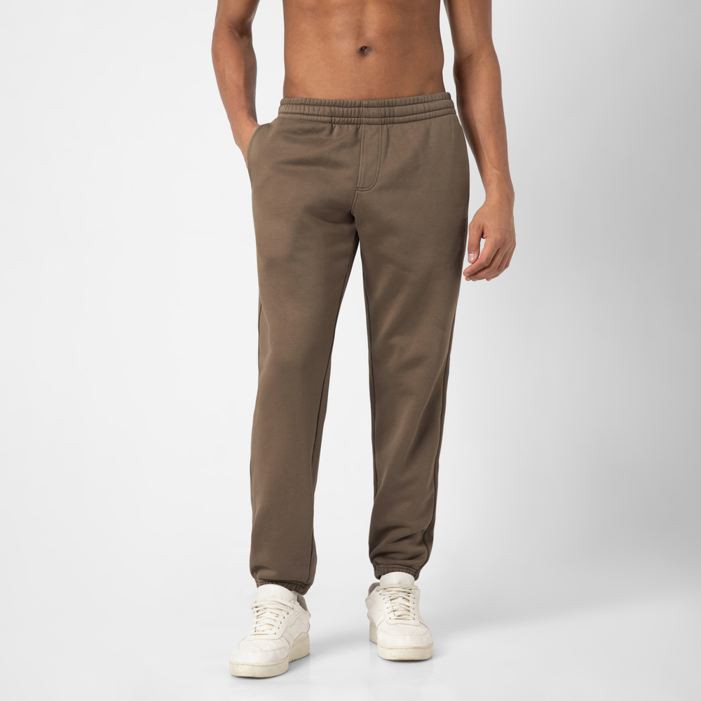 Wearing the Core Jogger in brown with an adjustable drawstring waist and white sneakers, a person stands against a plain light gray background with hands in pockets; only their lower body is visible.