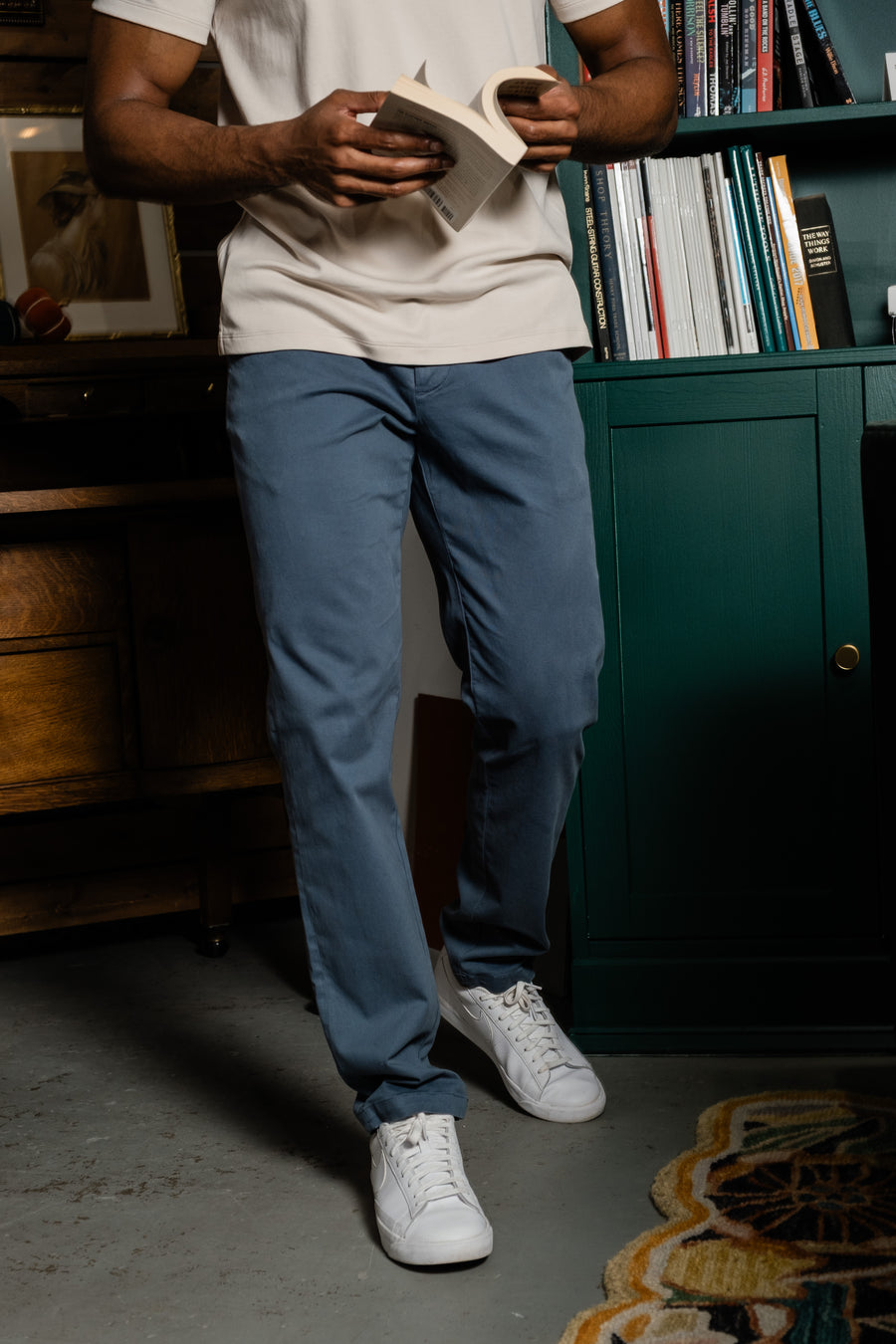 Person wearing a Supima Polo in Bone, Stretch Chino Pant in Flint, and white sneakers standing in a room with a bookshelf.