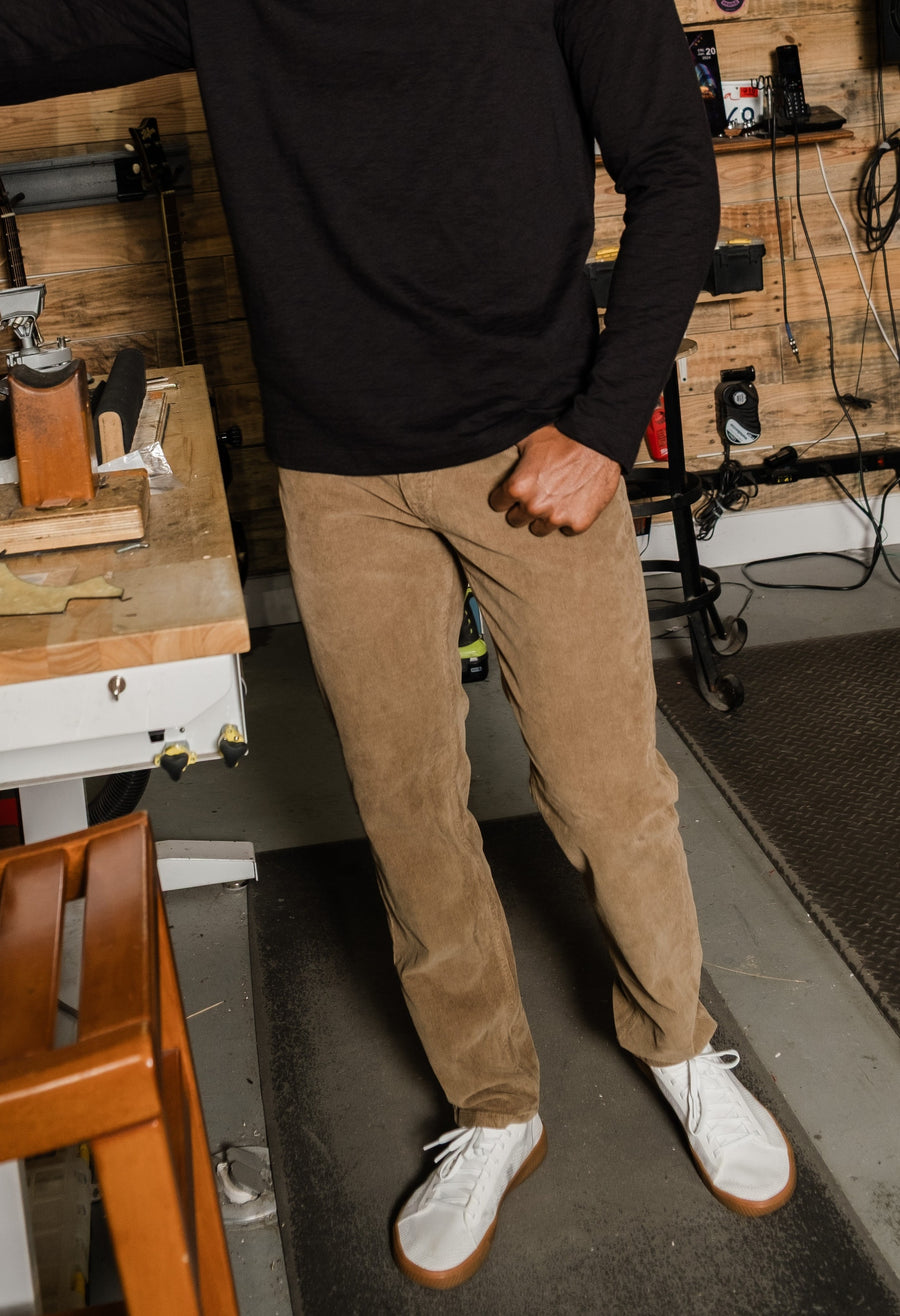 Person wearing a black Lounge Crewneck and Corduroy 5-Pocket Pant in Stone, and white shoes in a workshop setting.