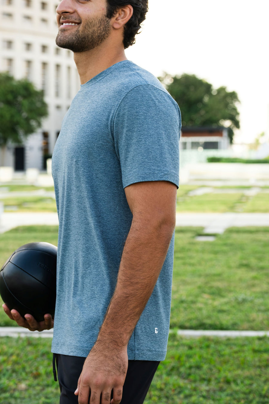 Man holding a medicine ball outdoors on a grassy area wearing Tech Tee in Dark Lagoon