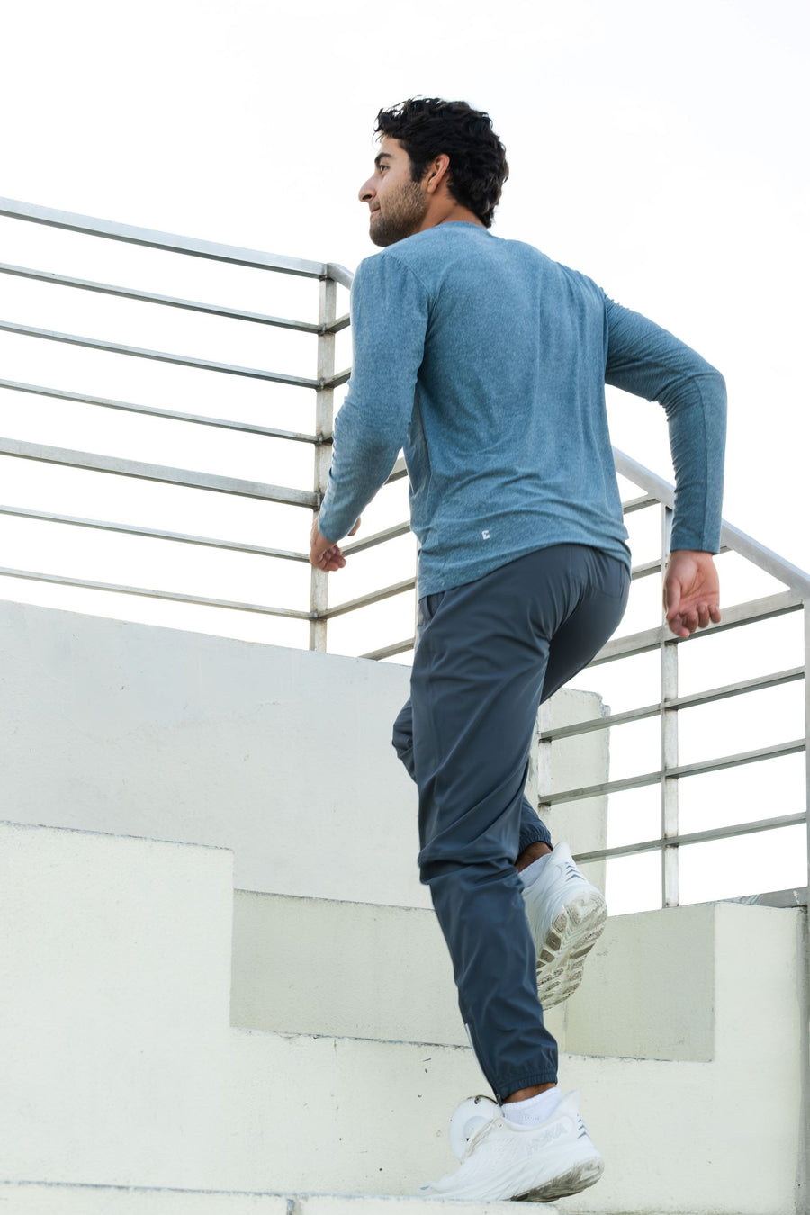 Man in Tech Long Sleeve Tee in Dark Lagoon and and Run Jogger in Coal walking on a white surface with a metal railing in the background.
