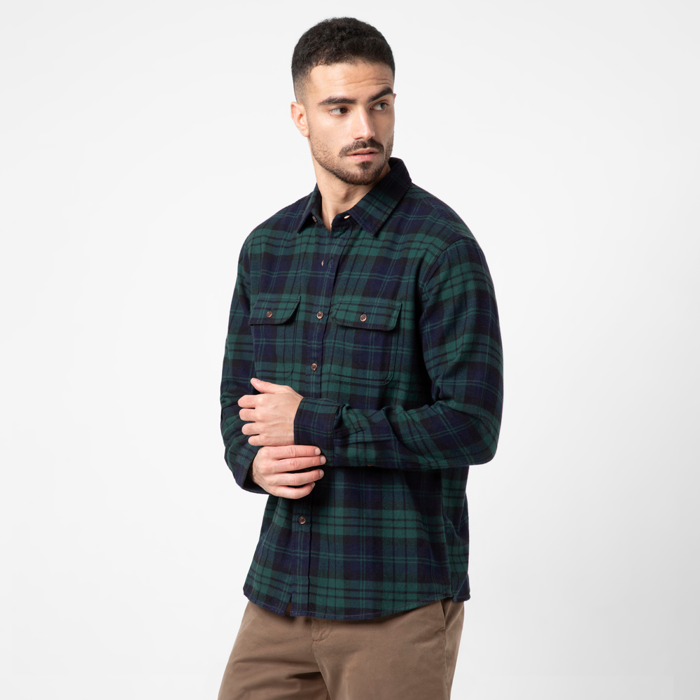 A man with short dark hair and a beard wears the Everyday Flannel Shirt in cozy green and navy yarn-dyed cotton, paired with light brown pants, standing against a plain white background and looking to his left.