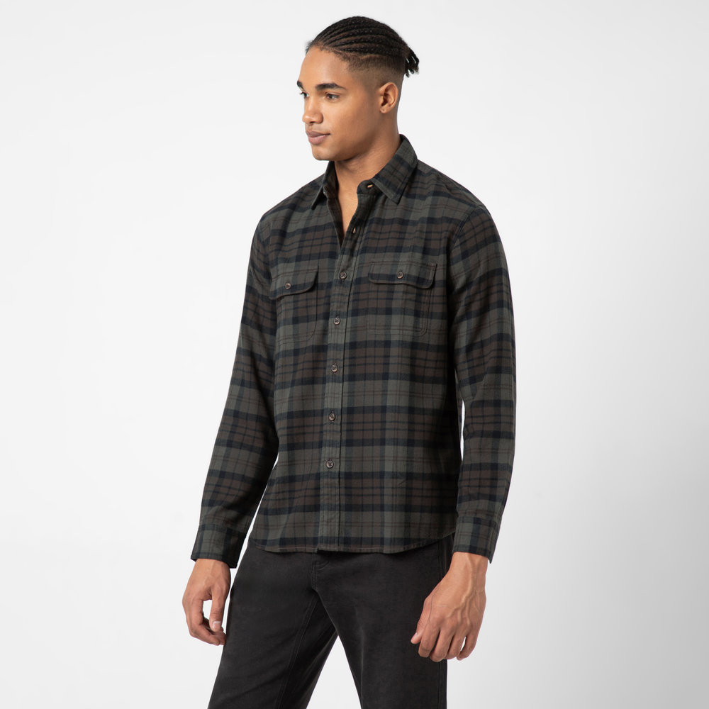 A man wears the Everyday Flannel Shirt—classic dark green and black plaid with two chest pockets—and black pants, standing against a plain white background and gazing slightly to the side.