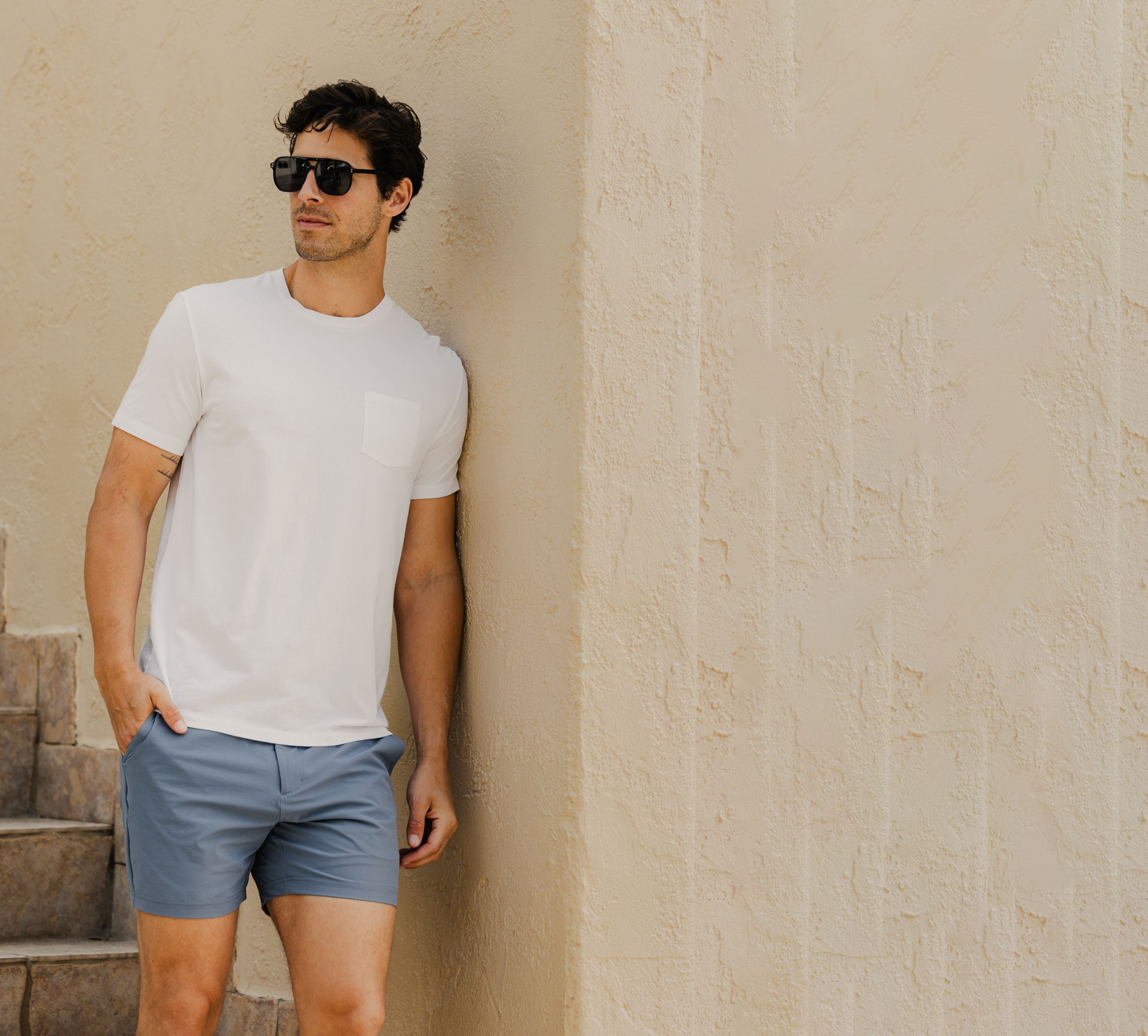 Man in Bamboo Pocket Tee White and Tour Short Flint leaning against a cream wall.