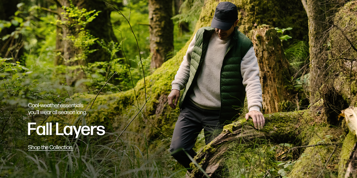 A man outside walking through a forest wearing the Sherpa Crewneck in Bone and the PrimaLoft Packable Vest in Pine Green.
