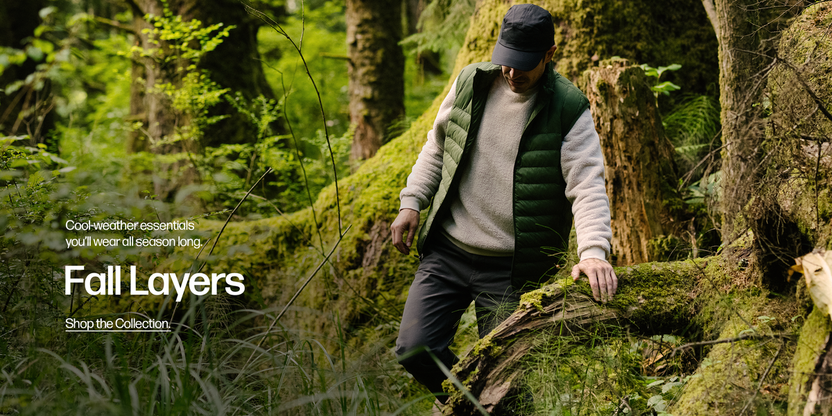 A man outside walking through a forest wearing the Sherpa Crewneck in Bone and the PrimaLoft Packable Vest in Pine Green.