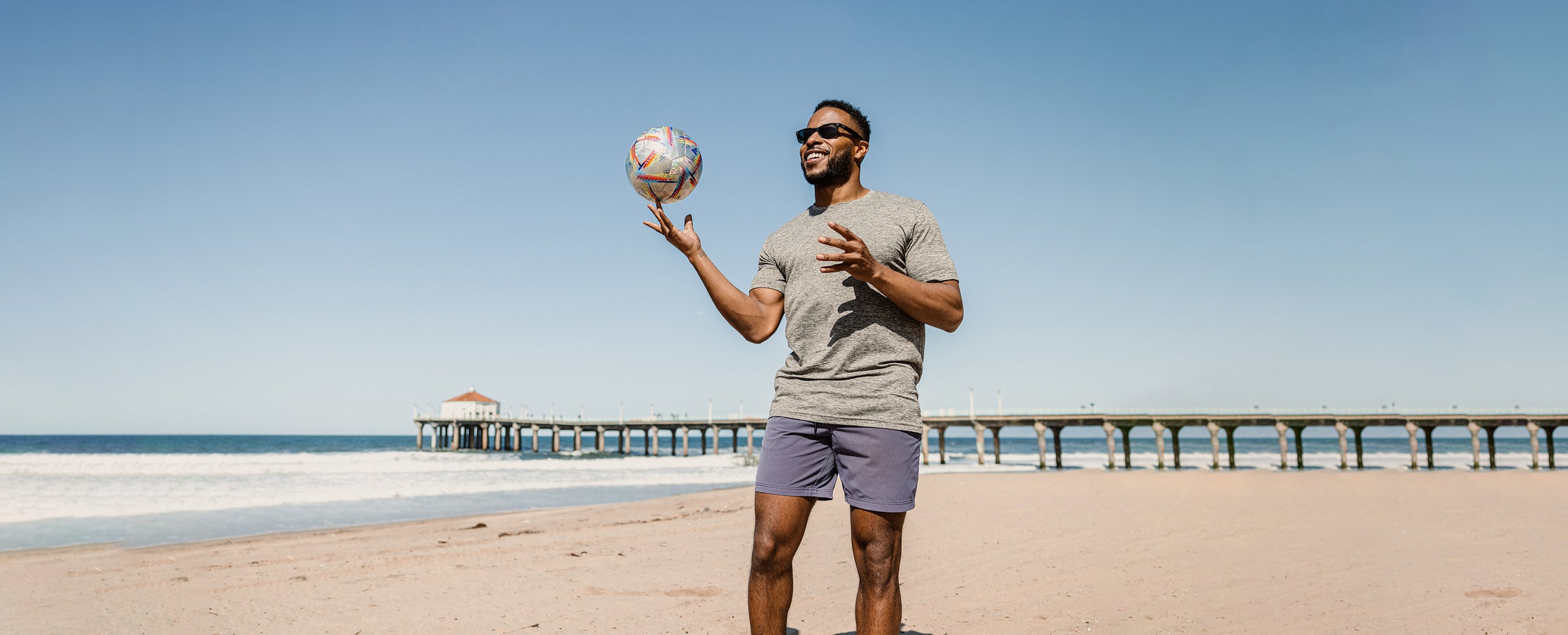 Man standing on beach spinning soccer ball on finger wearing Tech Tee Grey and Volley Short Purple