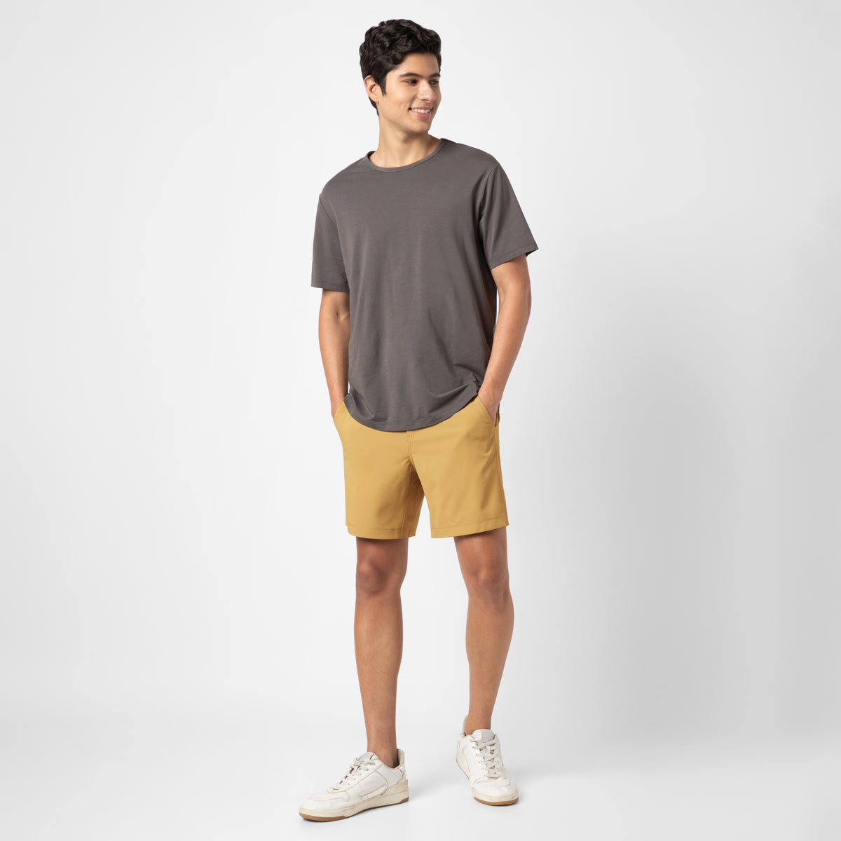 A person with short dark hair smiles against a plain white background, wearing a loose dark gray T-shirt, mustard yellow Hybrid Short with 4-way stretch for travel-ready performance, and white sneakers. Their hands are in their shorts pockets.