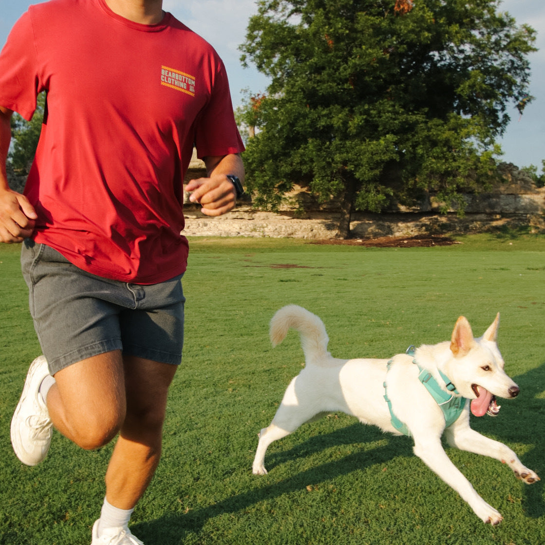 A man in a red Bearbottom t-shirt sprinting across a grass field in dark wash Stretch Denim Short, with a white dog running alongside