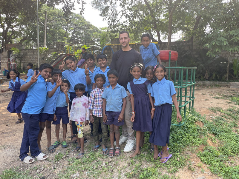 A man in a black t-shirt and light pants stands outdoors with a group of children wearing blue school uniforms, posing together in a rural outdoor setting with trees and a water tank visible in the background.