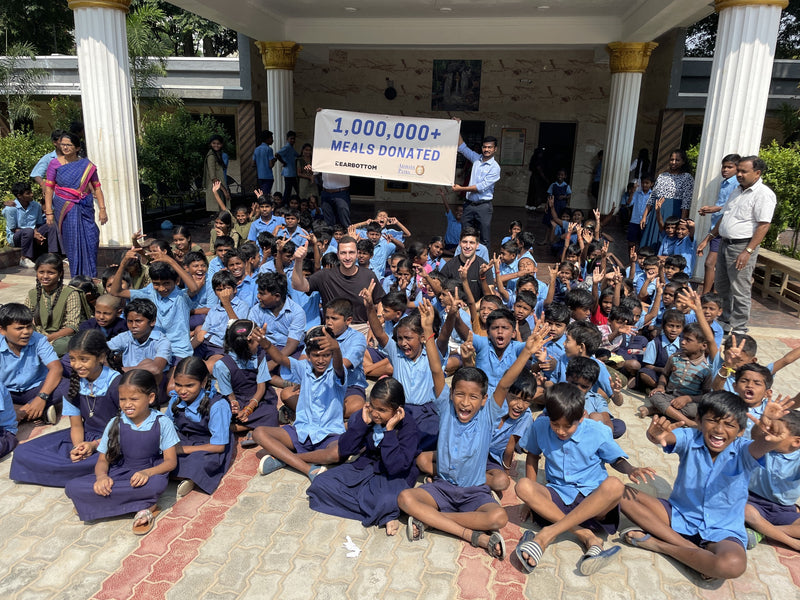 A large group of children in blue uniforms sits on a tiled courtyard floor under a building with white columns, holding and surrounding a banner that reads '1,000,000+ MEALS DONATED'. Several adults are standing around them, including a man holding the banner.