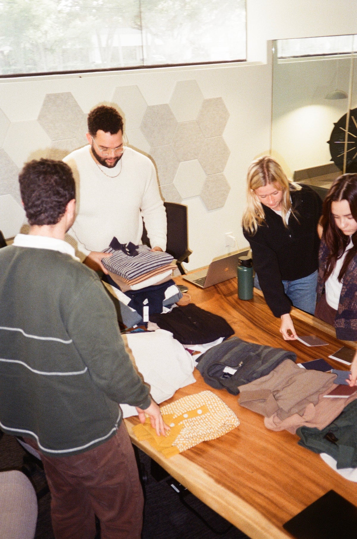 A group of four people gathered around a wooden table in an office setting, sorting through stacks of folded clothing and looking at a laptop. The room has a modern design with hexagonal wall panels and a large window.