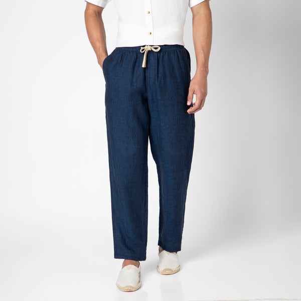 A person stands against a plain white background with hands in their pockets, wearing Linen Easy Pant in dark blue with a white short-sleeve button-up shirt and espadrilles, highlighting the pants’ breathable European linen fabric.