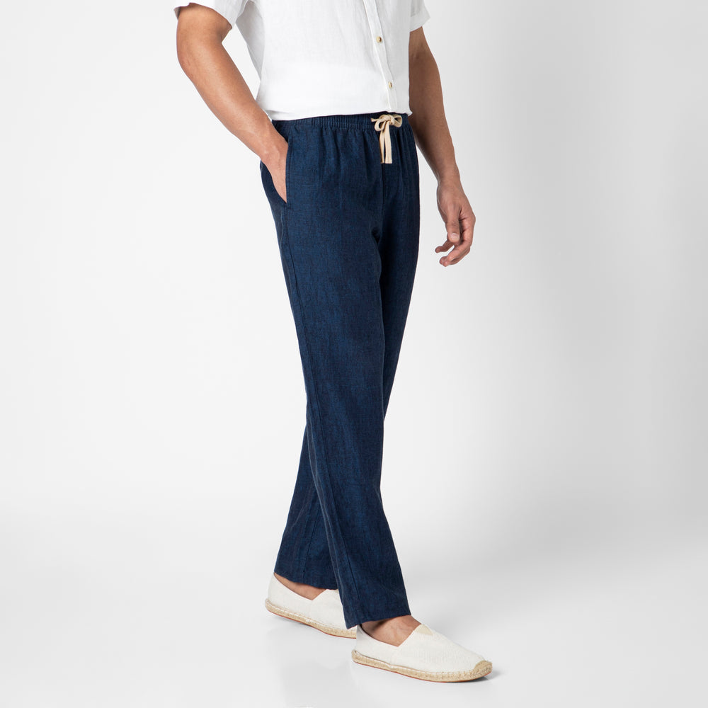 A person in a white short-sleeve shirt, navy blue Linen Easy Pant, and white slip-on shoes stands with one hand in their pocket against a plain white background.