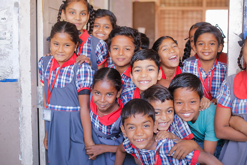 A group of smiling children wearing matching school uniforms with blue skirts and red-and-blue plaid shirts, standing together in a doorway.