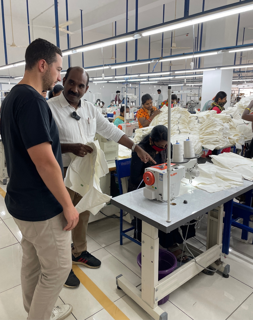 A Bearbottom team member and a production partner on a sewing floor, watching a worker at a sewing machine.