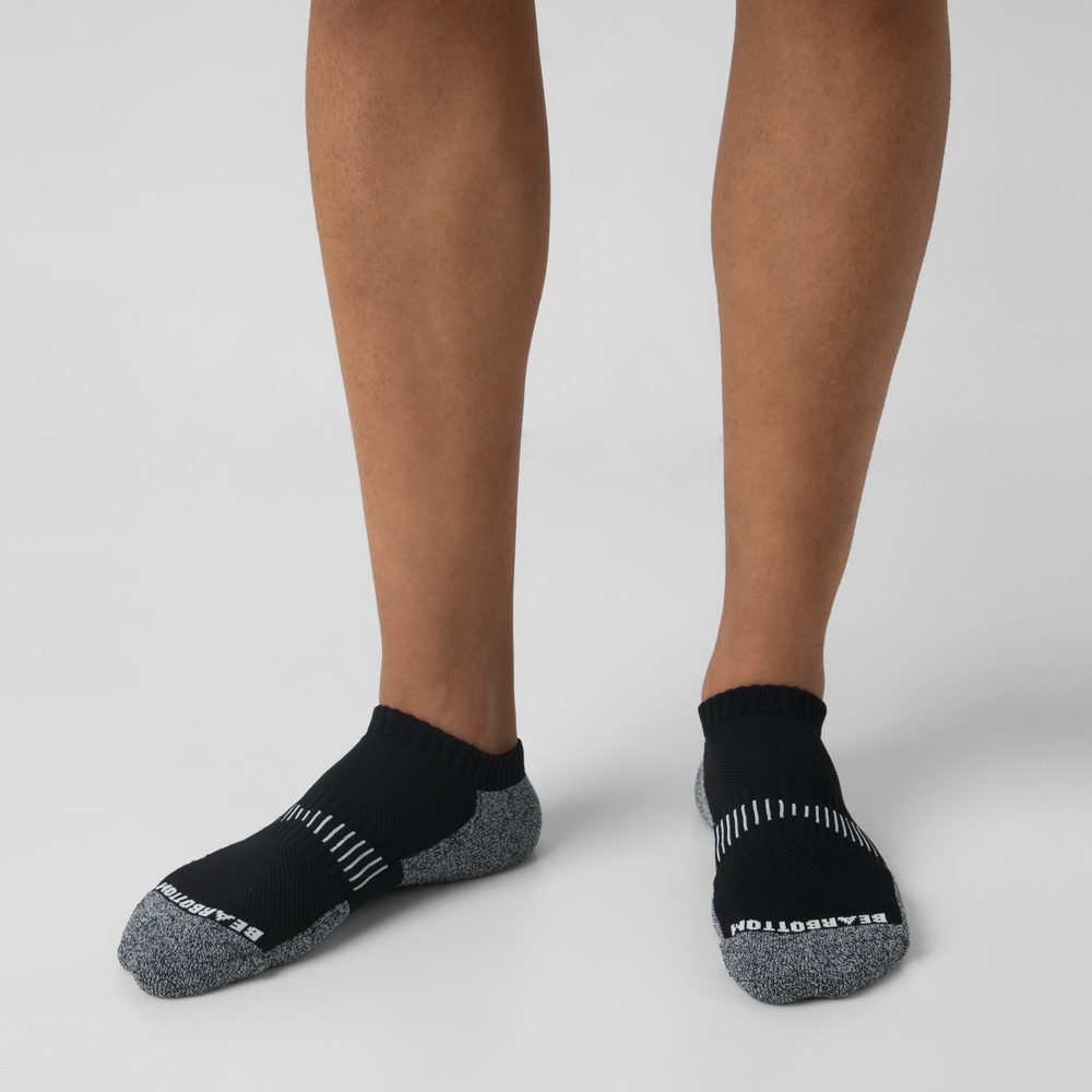 A person wearing black and gray Performance Sock with compression arch support stands on a light surface. BARTON is printed on the toe area.