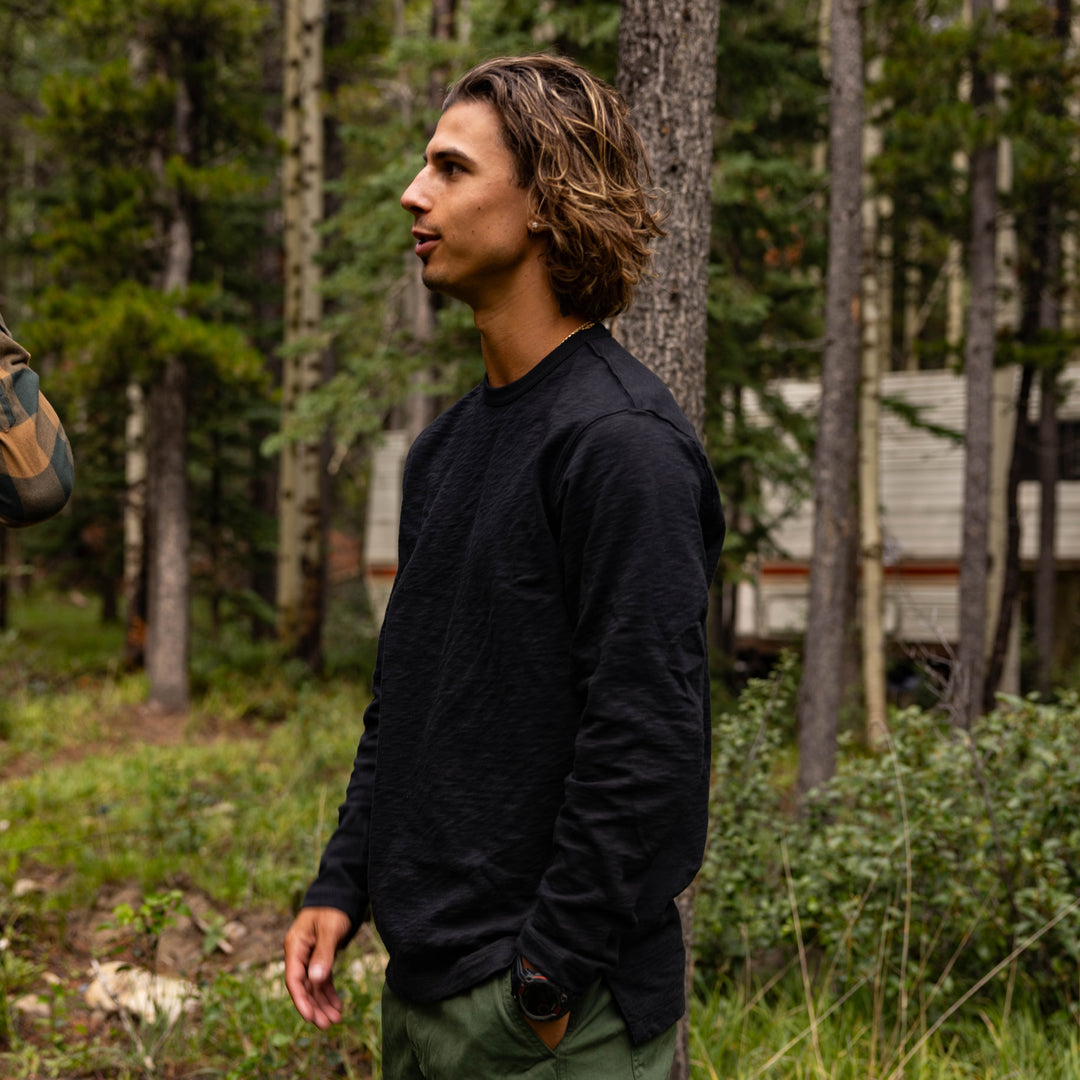 Man in a Rugby Crew Black standing in a forest with a camper van in the background